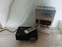 Black ProHT HD Mini digital projector with remote control on top and original product box in the background showing branding and model name.
