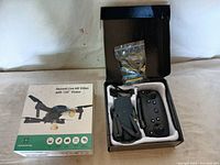 Opened box showing black foldable quadcopter drone, remote control, and small accessory bag inside styrofoam packaging.