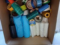 Box containing assorted rolls of adhesive tape, various colors and types, plastic bags in multiple colors (blue, white, black), and two green stretch wrap rolls