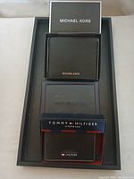 Three wallets displayed in a tray: one black Michael Kors wallet with gold logo, two Tommy Hilfiger wallets, one in packaging labeled 'trifold & valet'.