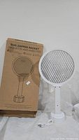 Bug zapper racket displayed next to its cardboard packaging box on a white surface. The racket is white with a circular mesh grid and a cylindrical stand below it. A USB cable is placed in front for charging.