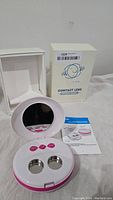 White and pink contact lens ultrasonic cleaner with open lid showing two stainless steel compartments, three pink buttons, instruction manual, and white box with graphics.
