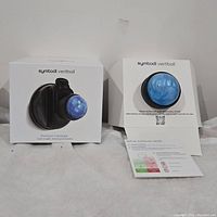 Two boxed Symbodi Vertiball precision massagers showing the product in packaging, highlighting the blue marbled massage ball and black mountable base.