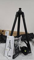 Photo of the black camera tripod set up, showing extendable legs, mounting plate, and included user manual and accessories