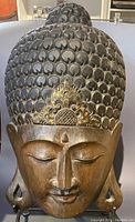 Full front view of the carved wooden Buddha mask showing the textured hair curls, facial features, and gold-accented crest on the forehead.