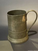 Front side view of pewter mug showing curved handle and horizontal bands, cylindrical shape with flared base.