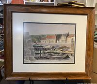 Frontal view of framed watercolour painting showing houses by the shore and stone wall with hills in background.