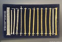 16 bracelets or bracelet bands displayed on black tray, mixture of silver-tone and gold-tone metal with various link and clasp styles.