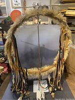 Full view of a circular frame wall hanging decorated with light brown fur, leather strips, beads, and feathers.