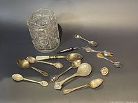 Glass jar filled with silver spoons and arranged on black surface with additional loose silver spoons and enamel pins.