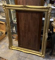 Full view of large rectangular gold frame resting upright against furniture on floor.