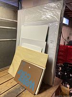 Group of art supplies including large plastic wrapped stretched canvas, several stacked watercolor papers and painting boards on wooden table.