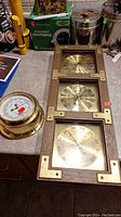 Both barometer sets displayed on a counter, showing the Springfield three-dial barometer with wooden frame and brass face, and the round brass Baker Barometer with white face.