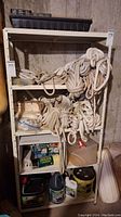 Wide view of white metal shelving unit containing several coiled boat lines on upper shelves and various bottles and boxes on lower shelves, taken in a basement.