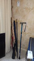 Photo showing three wooden walking sticks, two umbrellas (one black, one blue), and one blue trekking cane with gray handle.