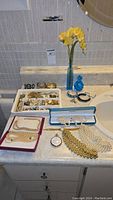 Overview of all items on countertop including jewelry box, pearls, watches, vase with flowers, and perfume bottle.