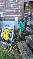 Two hose reels set outdoors against a brick wall, one gray SunEast hose reel with yellow hose, and one metal hose reel with green hose.