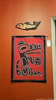 View of the red fabric Indigenous art piece with black, white and red totemic design bordered by black fabric with silver buttons, hanging on orange wall under the wood carved fish.