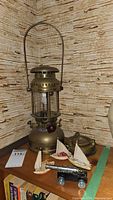 Optimus 1200 kerosene lamp, three ceramic sailboats, miniature cannon, and brass ashtray arranged on wooden shelf with books below