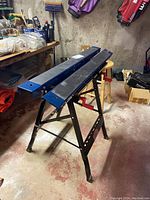 Two black Mastercraft foldable saw horses with blue top rails positioned side by side in basement workspace.
