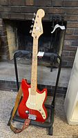 Full view of red Squier Bronco Bass guitar on black stand with strap attached in front of fireplace