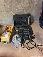 Electric drill with black body and orange trigger, assorted drill bits and hand tools organized inside black plastic tool kit case, and various small packaged parts in plastic bag.