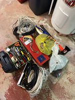 Wide view showing assorted electrical wires, a red toolbox, plastic bags with miscellaneous items, and a green metal toolbox with hand tools, on a concrete basement floor.