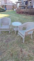 Two beige wicker chairs and small round wicker table with glass top on grass outdoors.