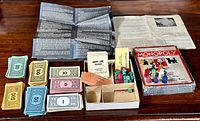Grouped items including paper money stacks, wooden player pieces in an open divided cardboard insert, instruction sheets, property cards, and the original box bottom