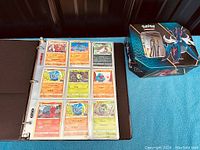 Photo showing an open black 3-ring binder with transparent plastic sleeves containing 12 Pokemon trading cards, including various Pokemon with Japanese text. A Pokemon tin box with artwork is positioned beside the binder.