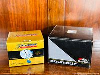 Boxes of Pflueger Centina and Abu Garcia Abumatic 275u reels side by side on wooden surface.