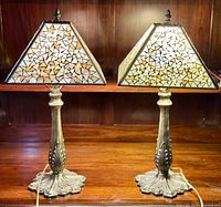 Pair of Tiffany style table lamps side by side on wooden surface with dark wood background, showing metal bases and square pyramid mosaic glass shades with teardrop pattern.