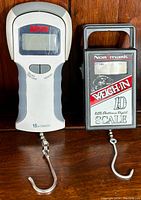 Two digital fish scales side-by-side on wooden surface, displaying front with digital displays and hook at bottom