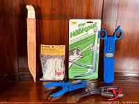 Photo of the complete fishing tools collection showing J Marttini Finland knife in leather sheath, Sampa knotting string in package, Hook-Kout hook removal tool in sealed pack, Shimano scissors with blue sheath, Berkley fish pliers with grip and red strap.
