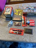 Wide view of Coca-Cola memorabilia including tin cans, coins, pens, golf ball, and matchbox car set on a gray felt surface