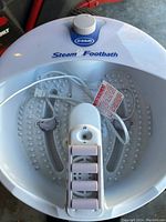 Top view showing the white plastic footbath basin with a raised footplate featuring massage nodes and a roller attachment inside. The power cord is coiled inside the basin.