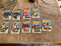Ten Hot Wheels cars in original sealed packaging, arranged on a beige cloth. Includes trucks, cars, a helicopter, and various designs.