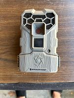 Front view of Stealth Cam trail camera showing camera lens, IR sensor, protective casing with textured bark-like design and honeycomb pattern around lens area.