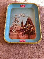 Front of a light blue early Coca-Cola tray featuring a birdhouse decorated with flowers and a Coke bottle, showing signs of wear and patina