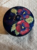 Top view of Moorcroft pin dish showing red and purple pansy flowers with green leaves on a blue background.