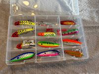 Clear plastic organizer box containing 11 assorted fishing lures with bright colors and treble hooks, arranged in shelves