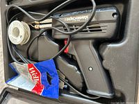 Top view of the black Weller Benchmark soldering gun with attached power cord, spool of solder wire, and packaged accessories laid in a molded black plastic case.