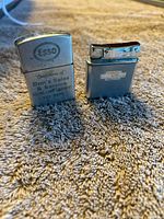 Photo showing two vintage metal cigarette lighters on a carpeted surface. Left lighter has Esso branding and advertising text, right lighter has Chevrolet logo and polished metal finish.