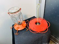 Two Perky-Pet No1 1958 hummingbird feeders, showing one assembled with glass jar and red feeding base with feeding ports and metal legs, and a separate red metal hanging ring attachment