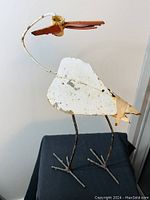 Front view of vintage cast iron garden bird sculpture with curved neck, white distressed paint, and rust-colored beak on metal stick legs.