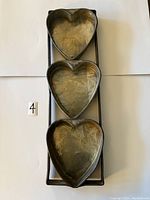 Three heart-shaped metal baking pans attached in a vertical line on a metal frame, visible scratches and patina.