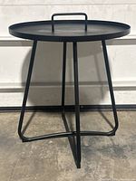 Front view of the black wrought iron outdoor patio table showing the round tray-style top with a handle and three angled legs.