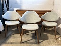 Four grey fabric mid-century modern style dining chairs with curved backrests and wooden legs shown from front in a warehouse setting. Seating areas show wear and fabric stains visible. All four chairs front facing.
