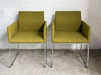 Front view of two green upholstered lounge chairs with chrome sled bases against a white cinderblock wall on concrete floor.