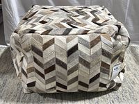 Full view of the square patchwork cowhide ottoman showing the top and side panels with a herringbone pattern in various shades of brown and cream.